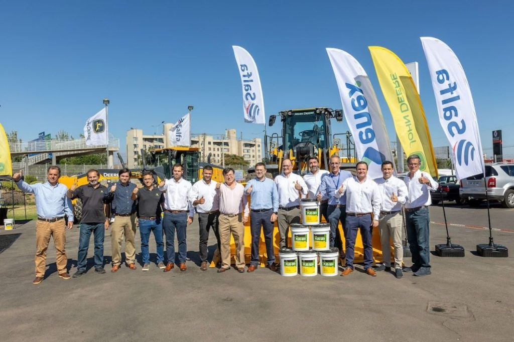 Grupo de personas posando en un evento de entrega de equipos de construcción John Deere, con banderas de Salfa y Yana, y baldes de lubricante Plus-50 II en primer plano.